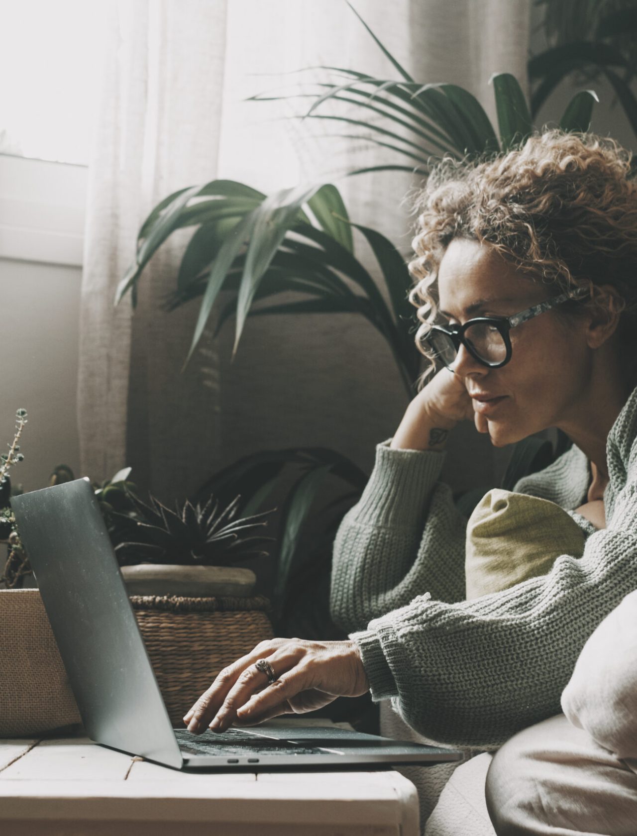 Blonde woman with curly hair using laptop sitting on sofa at home. 45 year old serious middle aged female looking at computer while surfing internet, shopping on website and relaxing on living room sofa.